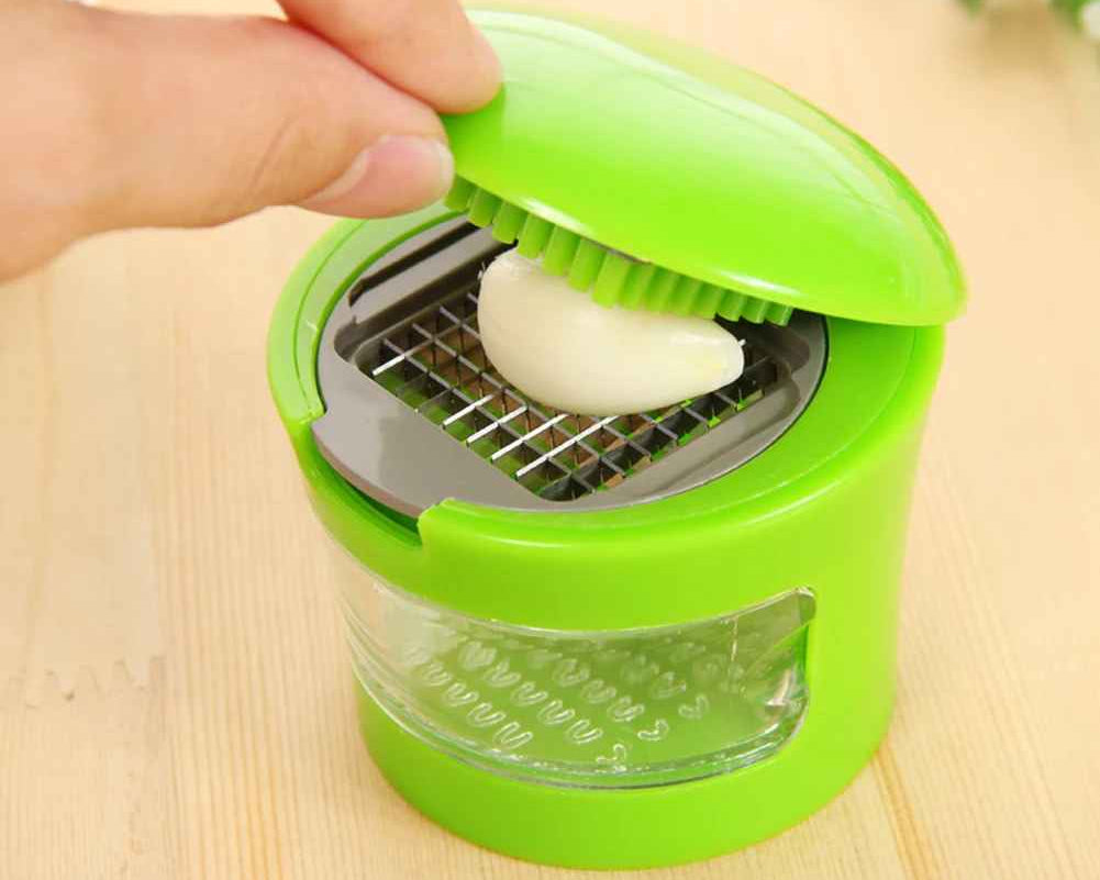 A hand is pressing down on the lid of a bright green manual garlic chopper, which is slicing a white garlic bulb over a metal grid. The chopper has a clear plastic container for the chopped pieces and sits on a light wooden surface.