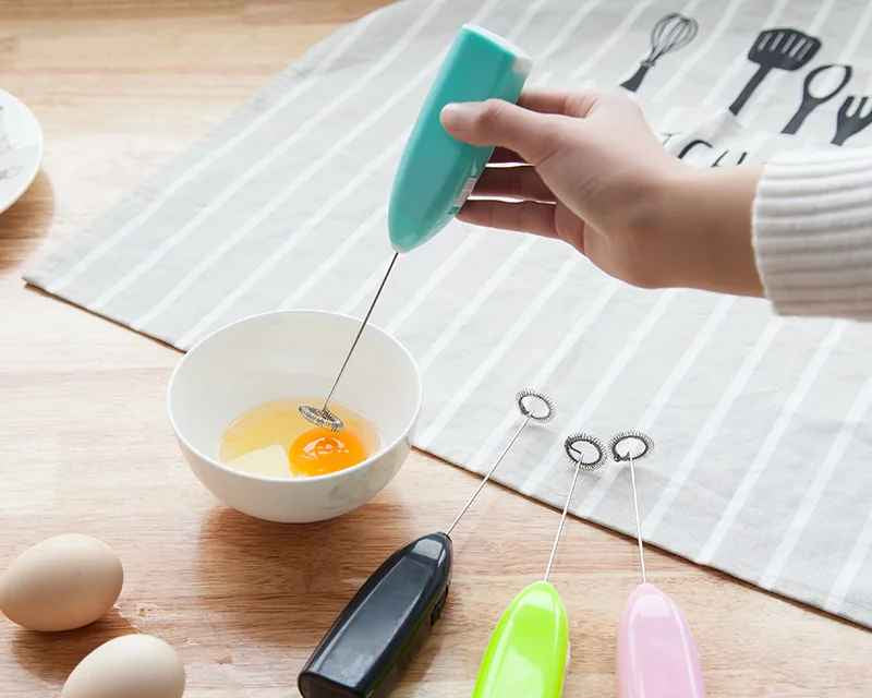 Hand using a small whisk to mix eggs in a bowl on a wooden surface with additional whisks nearby.