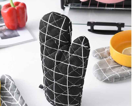 A black quilted oven mitt with a white grid pattern stands upright on a white countertop, surrounded by kitchen items including a red pepper, pink pot, yellow bowl, and other oven mitts.